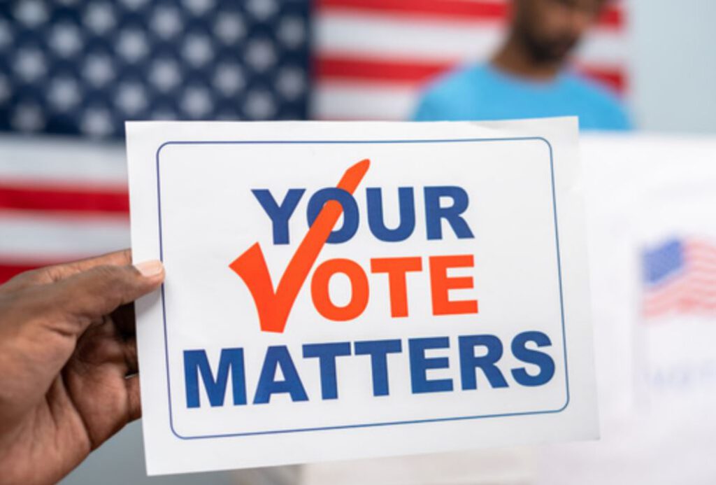 A hand holding a card that says, "Your vote matters.”  A U.S. flag and a person standing at a voting booth are out of focus in the background.